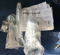 Photo of the door closer components including arm, brackets, mounting screws, and installation instructions on a blue carpet.