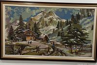 Full view of framed fabric art portraying a mountain scenery with pine trees and chalets under glass in a light gold frame.