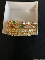 Photo of 4 gold-tone fashion bangles with clear and colored stones stacked inside a box, showing one bangle with a missing stone.