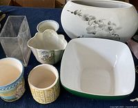All seven flower pots and one decorative bowl arranged on blue fabric, showing shapes, colors and designs of each item.