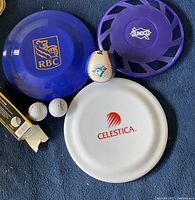 Frisbees with logos RBC, Sunoco, Celestica, baseball, golf balls, and sleeve on blue surface.