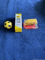 Three items displayed on a blue surface: a yellow and black toy soccer ball, a yellow and white boxed golf ball from Leon's, and a yellow toy truck sealed in packaging.