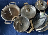 Top view of five cooking pots and fry pans with their glass lids arranged on a blue background.
