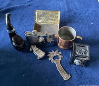 Items displayed on blue fabric showing pipe, flasks, tobacco tin, cup, toy car, bottle opener, and decorative safe figurine