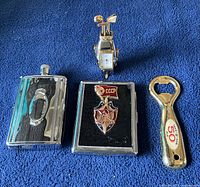 Three men's items on a blue fabric: stainless steel flask, vintage USSR cigarette case, and a metal bottle opener shaped like a beer bottle with '50' on it.