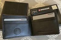 Three photos showing three black Wenger men's bifold wallets in black branded boxes with one box showing price and security label. Wallets are new, slim, and designed with card slots on the interior.