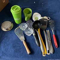 Collection of miscellaneous kitchen utensils and accessories laid on a blue surface, including spatulas, ladles, whisks, knives, measuring cups, and glass candle holders, emphasizing variety and condition.