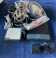 Photo showing two modems—a black Cisco modem and a grey RCA modem—along with multiple network cables in black and white, some packaged, and power adapters.