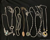 Photo showing 8 vintage necklaces laid out on a black surface with a mix of pendant styles including turquoise eagle, key pendant, pearls, locket, green stone pendant, and geometric silver/gold circular pendants.
