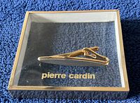 Top view of new Pierre Cardin tie clip inside original clear-lid box with gold trim and interior logo on black background