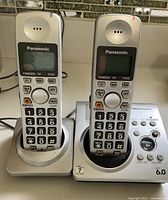 Panasonic cordless phone set with two silver handsets placed on charging bases, showing numeric keypad and function buttons.