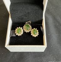 Green stone pendant and matching earrings shown inside a white jewelry box with black lining, highlighting the design and condition of the stones and settings.