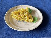Top view of oval ceramic plate with artificial yellow grapes and green leaves on blue fabric background.