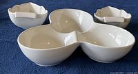 Front angled view of three white ceramic serving bowls on blue cloth: one large three-section bowl and two smaller leaf-shaped bowls.
