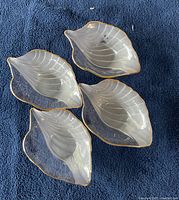 Top-down view of four glass serving bowls shaped like leaves with visible etched veins and gold trim around edges on a dark cloth background.