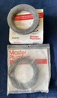 Two Master Plumber toilet bowl wax gaskets, one in sealed plastic bag, one partially out of box, placed on blue fabric surface.