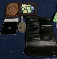 Lot contents showing two wall hanging frames, silver-plated brush, Millennium coin in box, and black makeup brush holder bag on a blue carpet.