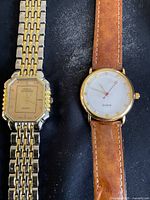 Two wristwatches side by side on a black surface, one with a gold-tone metal band and octagonal dial, the other with a brown leather strap and round white face.