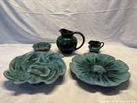 Full view of all five Blue Mountain Pottery pieces on a white cloth background: two pitchers, three dishes including daisy and swirl patterns, all glazed in green-toned ceramic.