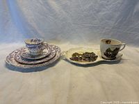 Shows the two British porcelain tea and biscuit sets, one with colorful Tudor house and people imagery, and the other with purple-blue floral designs.
