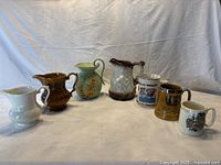 Seven British pottery and porcelain pitchers and mugs displayed together, showing variety in styles and colors including matte green, brown nylon glaze, and white porcelain.