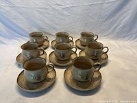 Eight Denby cup and saucer sets arranged in two rows on a white fabric background.