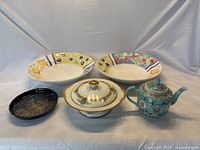 Wide view of the six porcelain and ceramic items showing the teapot, serving dish, two large fruit decorated bowls, and small round tray on a fabric backdrop.