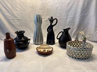 Photo of seven pottery and porcelain items including various vases, a pitcher, an olive dish, and a porcelain basket on white fabric background.
