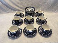 Eight blue and white Willow pattern teacups with matching saucers arranged on a table, with stack of cake/bread plates and fruit bowls visible behind.