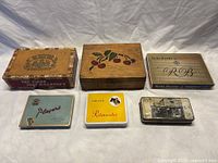 Six vintage cigar and tobacco boxes and tins arranged on a white background, showing various brand graphics and logos.