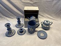 Full set on grey cloth background showing two tall candleholders, one short candleholder, mug in front of box, sugar bowl with lid, and ashtray, all in blue with white Wedgwood Jasperware style decorations.