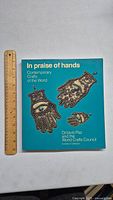 Front cover of 'In Praise of Hands' exhibition catalogue with artistic hand designs and ruler for scale.