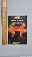 Front cover of the Close Encounters of the Third Kind book showing size next to a ruler for scale and cover art featuring spaceship and rock formation.