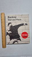 Front cover of the Banksy Wall and Piece art book featuring stencil graffiti art on a brick wall background and a ruler beside for scale.