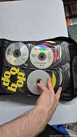 Hand holding open black CD binder showing some CDs stored inside, various music titles visible