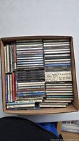 Top view of a cardboard box containing approximately 64 music CDs with various artists and genres.