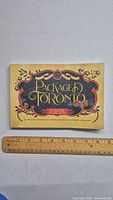 Front cover of 'Packaged Toronto' book showing stylized gold and red text and ornamental design on dark background, ruler beneath for scale.