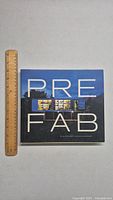 Front cover of the prefab architecture book with a modern illuminated prefab house shown and a ruler placed beside for size reference.