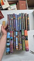 View of the Warriors book lot showing a selection of 17 Warriors series books by Erin Hunter stacked and arranged on a table with hands holding some of the books.