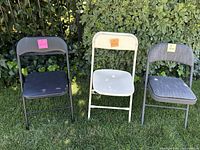 Three folding chairs side by side on grass - black, white, and gray colors. Each has a sticky note indicating quantity in the set.