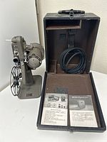 Film projector displayed beside open leatherette carrying case containing power cord and instruction booklet.