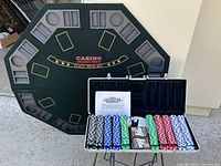 Octagonal gaming poker table with felt surface showing various player betting spots and a message in the center. Poker chips and accessories are displayed in a carrying case in front.
