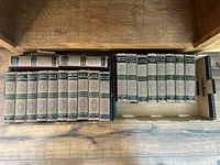 Top down view of 20 hardcover books of Charles Dickens works arranged in two rows in a wooden drawer or box. Titles and volumes clearly visible showing uniform brown hardcovers with gold lettering and decoration.