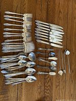 Image showing dinner forks, knives, tablespoons, teaspoons, small forks, and cocktail picks spread on wooden floor, all silverplated with ornate handles.