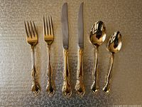8 pieces of Community Plate gold-plated flatware arranged on a textured surface including 2 forks, 2 knives, 1 soup spoon, and 1 teaspoon, all with detailed floral design on handles.