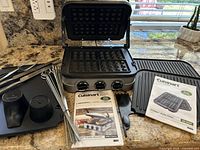 Cuisinart Griddler Gourmet open with waffle plates installed, grill plates and instruction booklets beside it, accessories including skewers and knives on countertop.