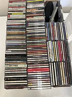 Full view of approximately 80-100 music CDs stacked in several columns with a clear plastic CD rack to the right side.