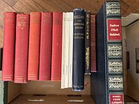 Overhead photo showing all nine books neatly arranged in a box. Includes several red cloth-bound books, three white paperbacks, a navy blue hardcover 'Ballads and Barrack-Room Ballads' by Rudyard Kipling, one dark book by Kipling, and a large navy hardcover 'Faulkner O'Neill Steinbeck' numbered as a Nobel Prize Library edition.