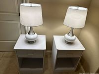 Photo showing two white table lamps with metallic bases on two white melamine nightstands, highlighting the drawer and shelf features of the nightstands.