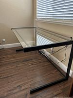 Angled side view showing glass top and black metal frame with U-shaped legs on wooden floor next to window with blinds.